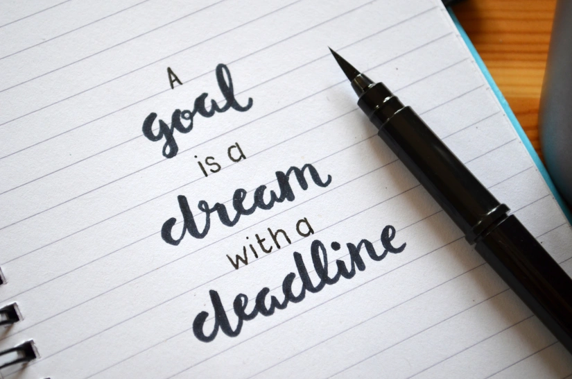 Image of a pen resting on lined paper that says A goal is a dream with a deadline